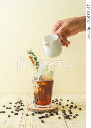 pouring milk in black coffee glass with ice cube, cinnamon and rosemary 91086147