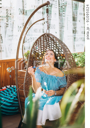 A young woman with bright makeup is sitting in a summer outdoor cafe in a hanging chair and blowing soap bubbles A young woman with bright makeup is sitting in a summer outdoor cafe in a hanging chair and blowing soap bubbles 91095024
