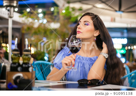 A young woman with bright makeup drinks wine from a glass in a street cafe, looks into the distance with a slight smile A young woman with bright makeup drinks wine from a glass in a street cafe, looks into the distance with a slight smile 91095138
