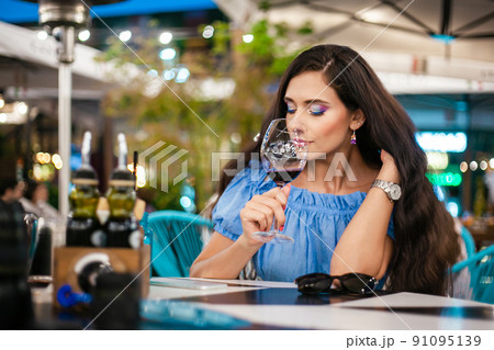 A young woman with bright makeup inhales the aroma of wine from a glass in a street cafe A young woman with bright makeup inhales the aroma of wine from a glass in a street cafe 91095139