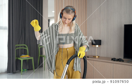 Young girl havig fun while cleaning floor with vacuum cleaner. Happy woman doing housework at home enjoy music wearing earphones. 91097650
