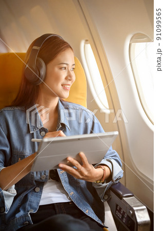 Charming asian woman passenger at window seat with headphones using portable tablet Charming asian woman passenger at window seat with headphones using portable tablet 91099565