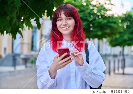 Outdoor portrait of teenage female with smartphone in hands looking at camera, on city 91101499