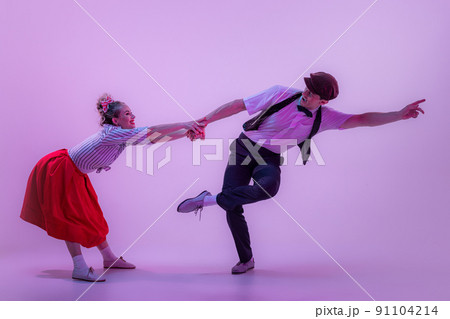 Studio shot of young man and woman in vintage retro style outfits dancing lindy hop isolated on lilac color background in neon light. 91104214