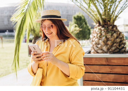 Pretty laughing girl with smartphone has a good time in summer weekend. Trendy lady with brown hair wears straw hat using phone outdoors. Phone Communication. Urban lifestyle concept. Traveler Pretty laughing girl with smartphone has a good time in summer weekend. Trendy lady with brown hair wears straw hat using phone outdoors. Phone Communication. Urban lifestyle concept. Traveler 91104431
