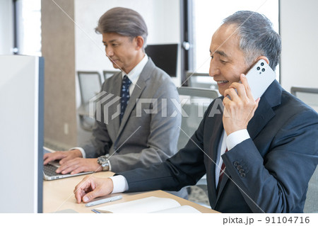 オフィスでスマホで会話をするシニア男性とパソコン作業をするシニア男性 91104716