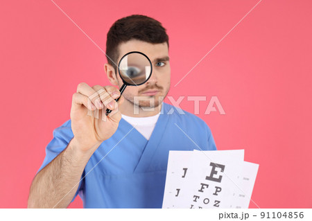 Male nurse holds magnifier and vision test on pink background Male nurse holds magnifier and vision test on pink background 91104856