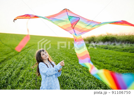 Little happy girl running with kite in hands on green wheat field. Large colored rainbow kite with long tail. Little happy girl running with kite in hands on green wheat field. Large colored rainbow kite with long tail. 91106750
