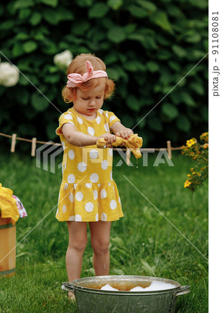 Lttle girl in a yellow dress hangs clean clothes on a clothesline. 91108081