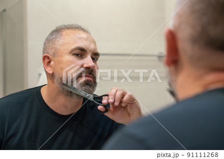 Portrait of handsome mature man trimming his beard himself standing in bathroom 91111268
