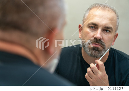 Middle-aged handsome man brushing his beard in the morning in bathroom looking in mirror Middle-aged handsome man brushing his beard in the morning in bathroom looking in mirror 91111274