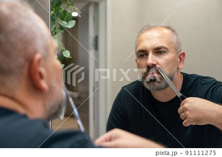 Portrait of Middle-aged handsome man cutting his beard with scissors. Selfcare during quarantine isolation 91111275