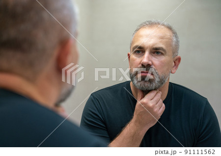 Middle-aged handsome man looking in mirror in bathroom touching his beard thinking of cutting it off Middle-aged handsome man looking in mirror in bathroom touching his beard thinking of cutting it off 91111562