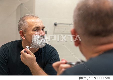 Good-looking mature man with grey hair trying to cut off his beard himself in a bathroom 91111786