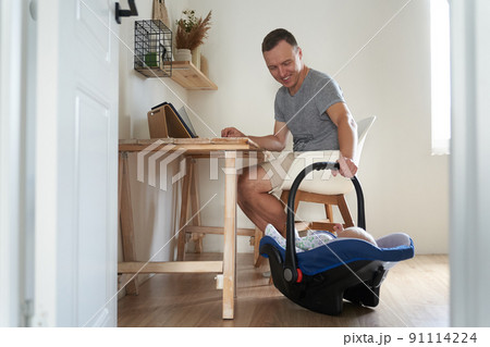 A handsome freelance man works on a modern laptop. A young father works at home and cradles his little son in a cradle. High quality photo A handsome freelance man works on a modern laptop. A young father works at home and cradles his little son in a cradle. High quality photo 91114224
