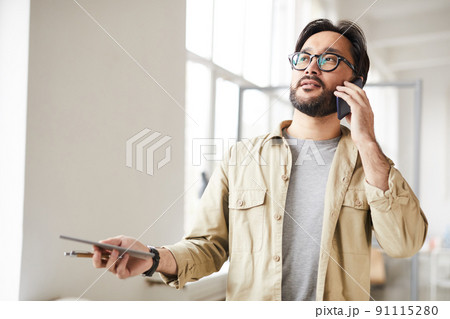 Content confident young Asian entrepreneur in shirt standing in office and holding tablet while discussing project by phone in office 91115280