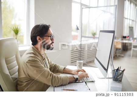 Concentrated hipster Asian art director with beard sitting at table and typing on computer keyboard while examining project design Concentrated hipster Asian art director with beard sitting at table and typing on computer keyboard while examining project design 91115303