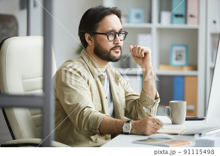Serious pensive young Asian manager with beard sitting at desk and using modern computer while analyzing ideas 91115498
