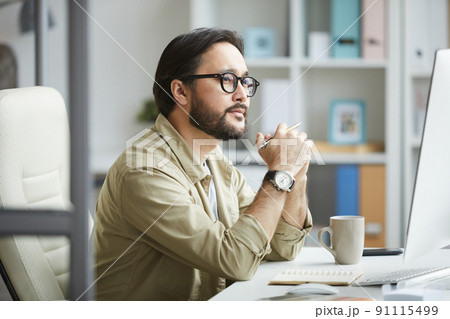 Serious pensive young Asian man with beard and mustache sitting at desk in office and looking at computer monitor while analyzing web design 91115499