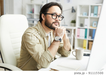 Portrait of content pensive hipster young Asian designer with beard sitting at table and leaning head on hands while finding inspiration 91115500