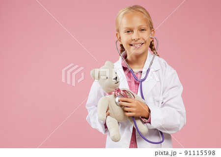 Portrait of happy beautiful little girl with blond braids wearing white coat using stethoscope while examining toy bear 91115598