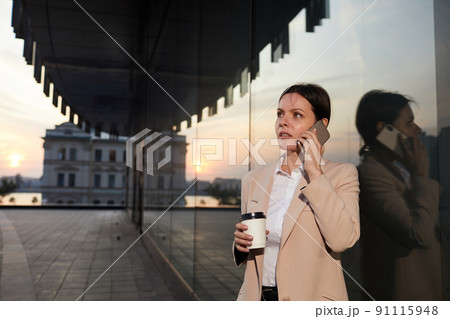 Serious pensive brunette woman standing by contemporary building and calling colleague on phone to discuss project Serious pensive brunette woman standing by contemporary building and calling colleague on phone to discuss project 91115948