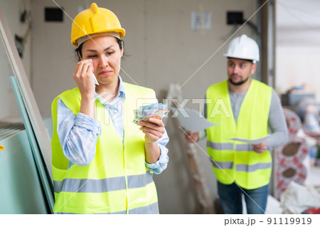 Crying female builder standing at construction site with banknotes in hands Crying female builder standing at construction site with banknotes in hands 91119919