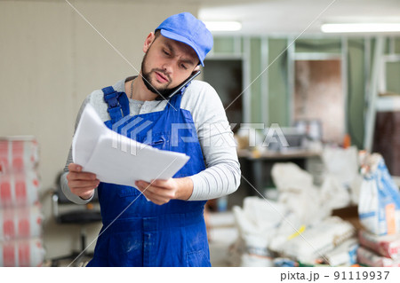 Master in blue overalls talking on mobile phone, discussing documents in renovated building Master in blue overalls talking on mobile phone, discussing documents in renovated building 91119937