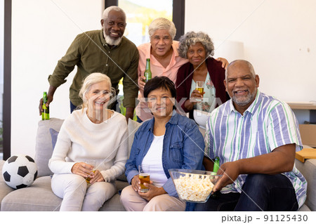 Portrait of cheerful multiracial senior friends with beer and popcorn enjoying match in nursing home 91125430
