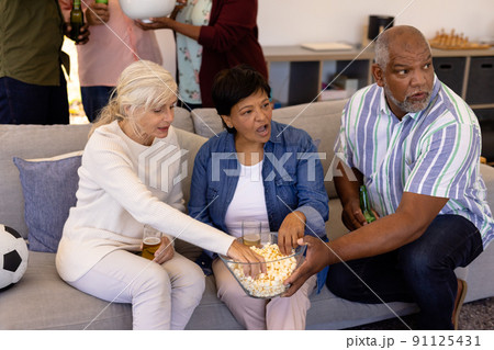 Multiracial senior friends with beer having popcorn while watching soccer match in nursing home 91125431