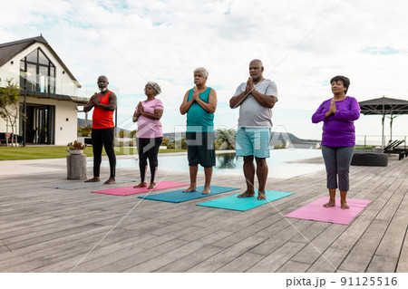 Multiracial senior friends meditating in prayer position at poolside against sky at nursing home 91125516