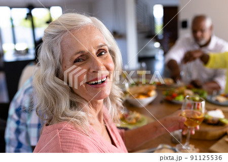 Portrait of senior multiracial woman enjoying wine while having lunch with friends at dining table 91125536