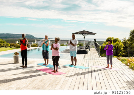 Multiracial seniors meditating while standing on hardwood floor at poolside against sky in summer Multiracial seniors meditating while standing on hardwood floor at poolside against sky in summer 91125556
