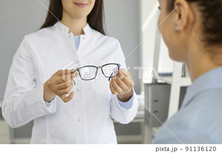Woman oculist or doctor ophthalmologist in white coat holds out glasses to vision to patient 91136120