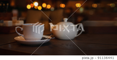 White cup with teapot. Pot standing on saucer in soft focus on naturally blurred background. Coffee, tea house, bokeh lights. The concept of a cozy pastime, tea ceremony White cup with teapot. Pot standing on saucer in soft focus on naturally blurred background. Coffee, tea house, bokeh lights. The concept of a cozy pastime, tea ceremony 91136441