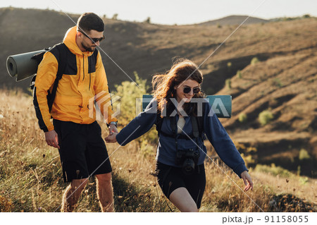 Cheerful Woman with Her Boyfriend Holding by Hands While Hiking on the Hill During Sunset, Young Couple Enjoy Their Trip Cheerful Woman with Her Boyfriend Holding by Hands While Hiking on the Hill During Sunset, Young Couple Enjoy Their Trip 91158055