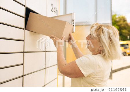 50s caucasian woman getting parcel from cell of automatic post terminal outdoors. 50s caucasian woman getting parcel from cell of automatic post terminal outdoors. 91162341