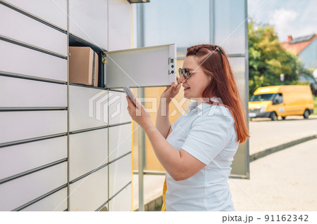 Young caucasian woman getting parcel from cell of automatic post terminal outdoors. Young caucasian woman getting parcel from cell of automatic post terminal outdoors. 91162342