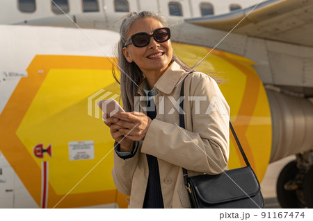 Cheerful woman passenger in sunglasses using phone standing outdoors at airport near plane Cheerful woman passenger in sunglasses using phone standing outdoors at airport near plane 91167474