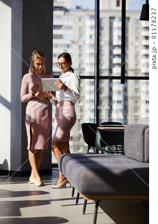 Serious thoughtful young businesswomen in pink clothes standing in modern hall and using tablet while discussing business Serious thoughtful young businesswomen in pink clothes standing in modern hall and using tablet while discussing business 91178237