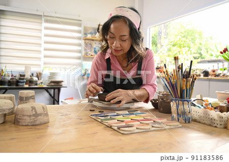 Smiling mature woman decorating and shaping pottery before baking in workshop. Indoors lifestyle activity, handicraft, hobbies concept 91183586