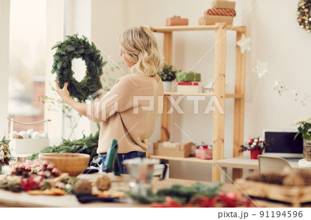 Blonde young woman in sweater standing in own workshop and looking at green Christmas wreath Blonde young woman in sweater standing in own workshop and looking at green Christmas wreath 91194596