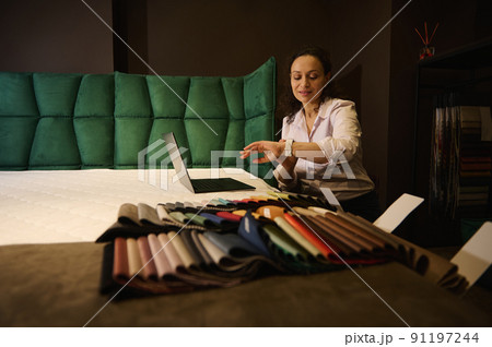 Creative interior designer checking time on her watch while working on laptop on orthopedic mattress next to laid out colorful fabric samples in home design studio 91197244