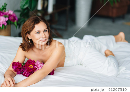 gentle brunette topless woman with a bouquet of peonies on the bed. 91207510