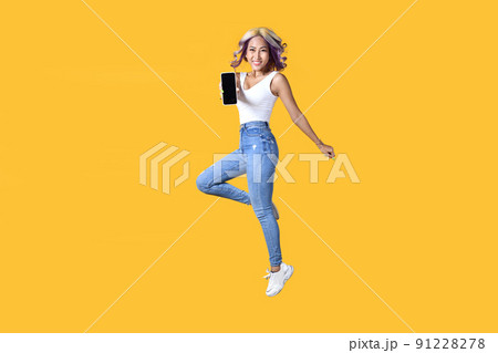 Full length portrait of young Asian woman holding smartphone jumping on yellow background 91228278