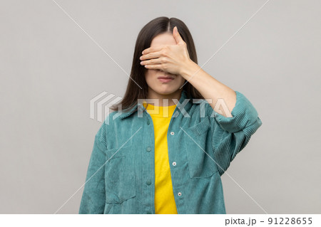 Don't want to look at this. Portrait of shy confused woman covering eyes with hand, feeling shamed and scared to watch, wearing casual style jacket. Indoor studio shot isolated on gray background. 91228655