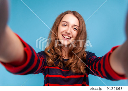 Beautiful woman wearing striped casual style sweater, making selfie POV, having positive expression, being in good mood, point of view. Indoor studio shot isolated on blue background. 91228684