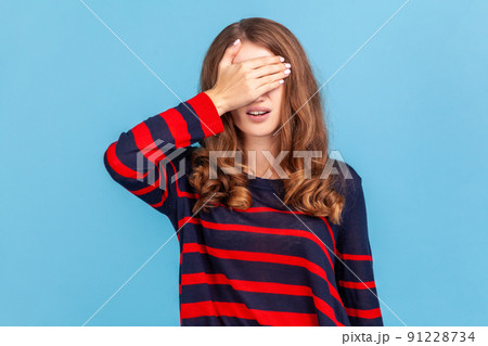 Don't want to look. Portrait of woman wearing striped casual style sweater standing covering eyes with hand, does not want see at something shameful. Indoor studio shot isolated on blue background. Don't want to look. Portrait of woman wearing striped casual style sweater standing covering eyes with hand, does not want see at something shameful. Indoor studio shot isolated on blue background. 91228734