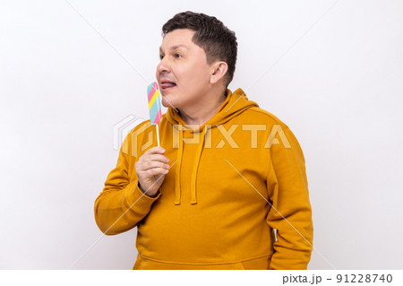 Funny man sticking out tongue licking ice cream, tasting and enjoying delicious flavor dessert, sweet confection, wearing urban style hoodie. Indoor studio shot isolated on white background. 91228740