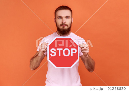 Portrait of serious bossy bearded man holding red stop sign looking at camera with serious expression, wearing pink T-shirt. Indoor studio shot isolated on orange background. Portrait of serious bossy bearded man holding red stop sign looking at camera with serious expression, wearing pink T-shirt. Indoor studio shot isolated on orange background. 91228820